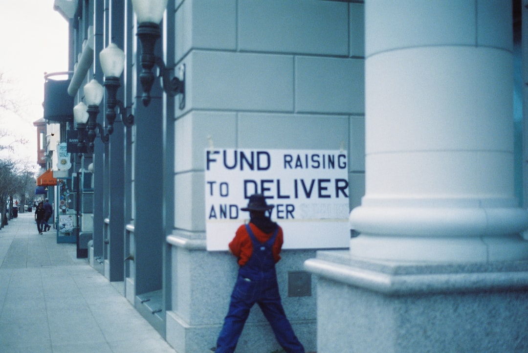 man in blue jacket and blue denim jeans standing beside white concrete wall